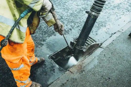 Workers cleaning a storm drain using pressure washer on a rainy day in an urban area